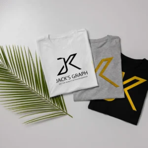 three folded t shirts—white, gray, and black—each with a stylized jk logo on the front, are arranged side by side next to a green palm leaf on a light gray background.